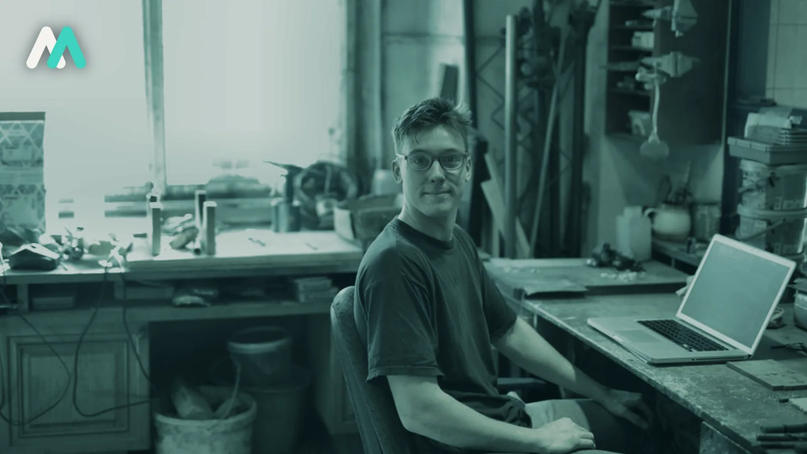 A founder working at a bench in a small workshop, laptop open beside the tools.