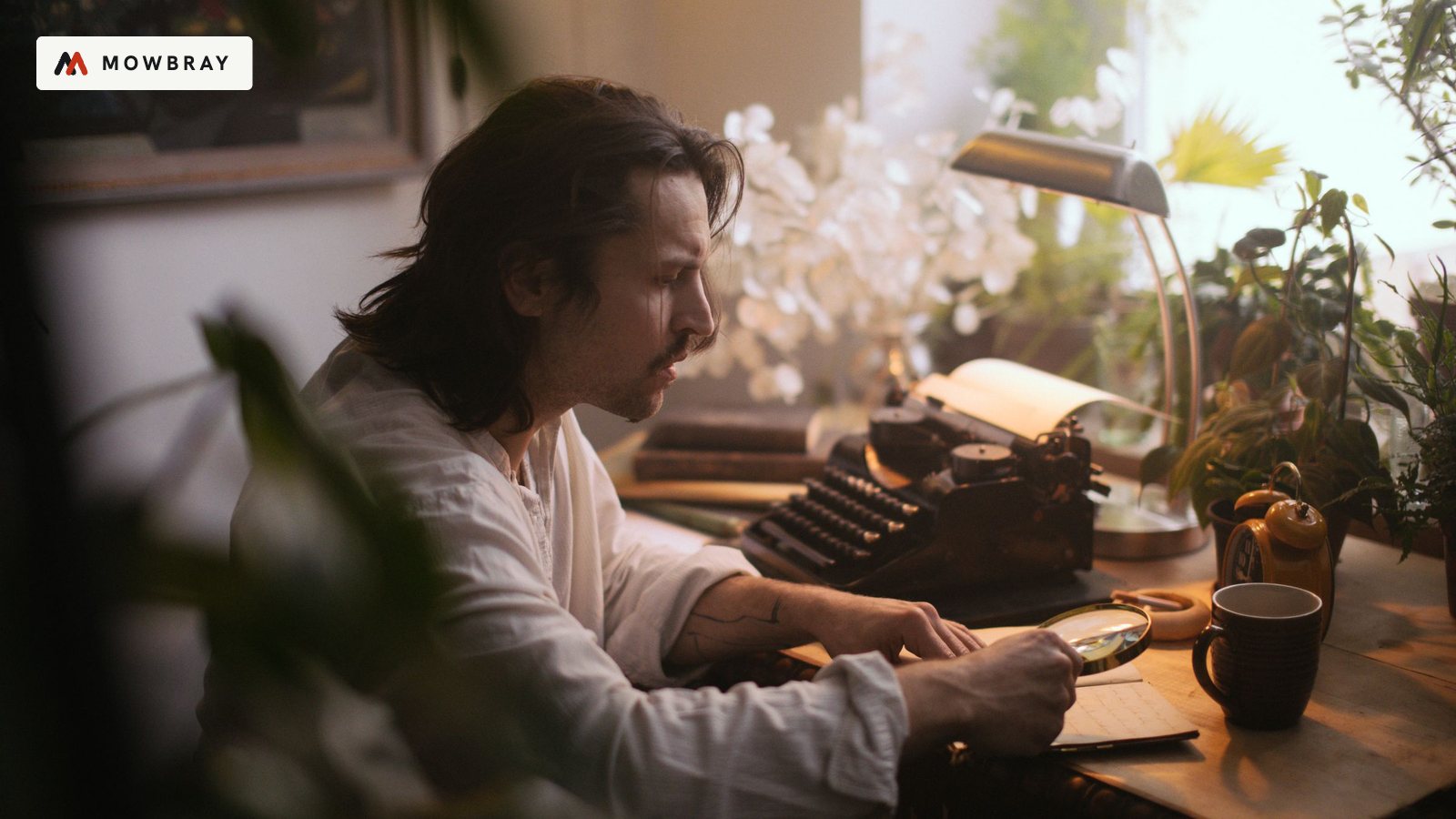 A man typing on a typewriter at a desk by the window