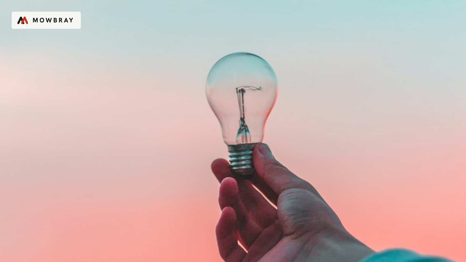 A hand holding up a light bulb against a pink sky