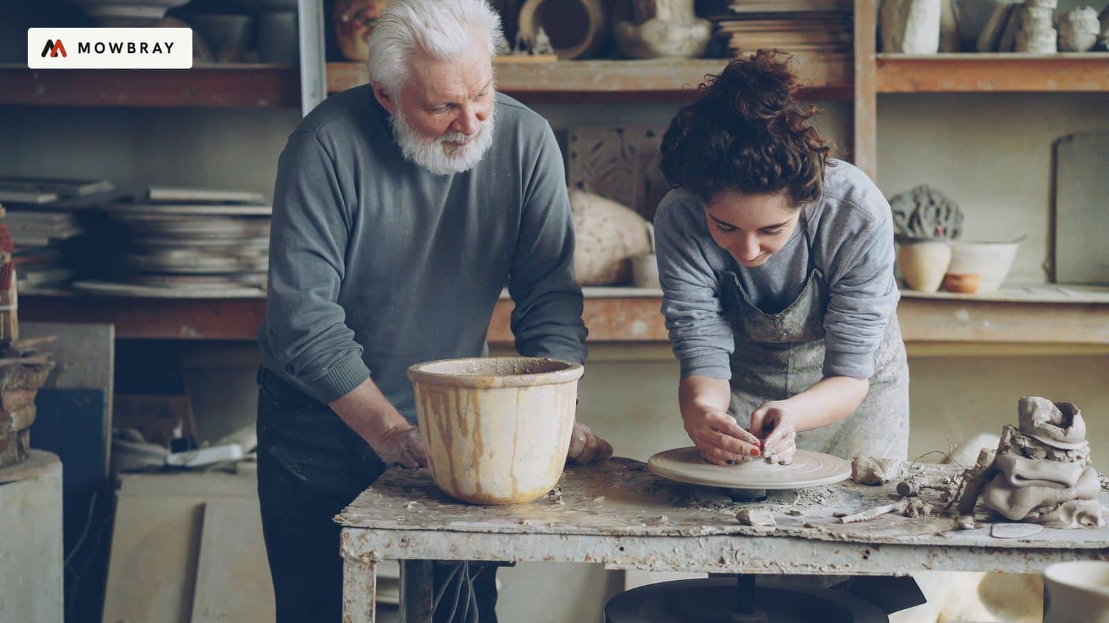 An older potter guiding a younger maker at a workbench